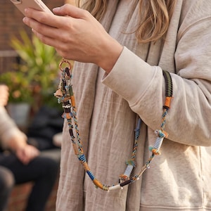 May include: A phone lanyard featuring a braided cord in shades of blue, orange, and green, accented with small fabric tassels, striped cylindrical beads, and dark brown wooden spacers, attached to a circular gold-toned metal ring connector.
