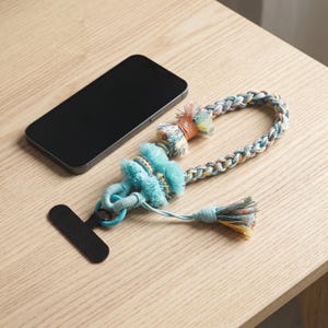May include: A black smartphone next to a braided phone wrist strap featuring turquoise, orange, and yellow threads. The strap includes decorative fringe accents, a solid turquoise loop, and a black plastic attachment tab for a phone case.