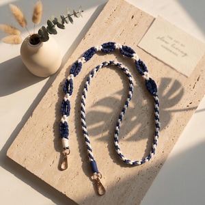 May include: A macrame lanyard featuring a navy blue and white cord design. The strap combines a braided section with a series of knotted oval loops. Two gold-toned metal swivel lobster clasps are attached to the ends of the cord.