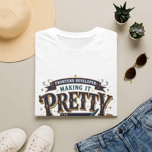 May include: A white t-shirt featuring a dark blue and gold graphic design with the text "FRONTEND DEVELOPER: MAKING IT PRETTY" in a decorative font. The design includes stylised flourishes, stars, and a small icon of a computer screen.