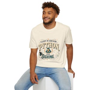 May include: A cream-coloured short-sleeve t-shirt featuring a graphic print with the text "FLUENT IN SARCASM, PYTHON AND SIGHING". The design includes a coiled green snake, a small coffee mug, and decorative flourishes in dark teal and gold.
