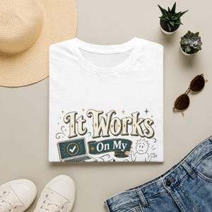 Könnte beinhalten: Ein weißes T-Shirt mit einem Grafikdruck, der den Text "It Works On My" über einem blaugrünen Banner und einem Laptop-Symbol mit Häkchen zeigt. Rechts daneben zuckt eine einfache Strichmännchen-Figur mit den Schultern, umgeben von kleinen Sternen.