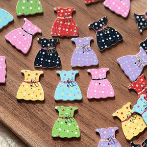 Könnte beinhalten: Eine Sammlung kleiner, flacher, kleiderförmiger Knöpfe in verschiedenen Farben wie Rot, Gelb, Grün, Blau, Rosa, Lila und Schwarz. Jeder Knopf ist mit einem Punktmuster und einem aufgemalten Taillendetail mit einer kleinen Schleife versehen.