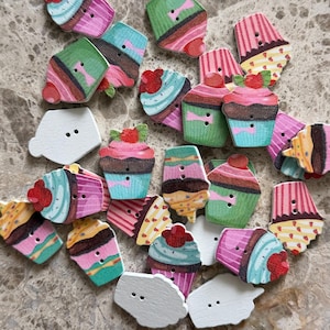 Könnte beinhalten: Eine Sammlung flacher Holzknöpfe mit zwei Löchern in Cupcake-Form. Die Knöpfe zeigen bunte Drucke von Cupcakes mit verschiedenen Glasurfarben wie Pink, Blau, Grün und Rot sowie einige schlichte, unbemalte weiße Knöpfe.