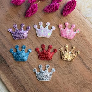Könnte beinhalten: Sieben kronenförmige Aufnäher, die mit kleinen, reflektierenden Pailletten bedeckt sind. Die Farben umfassen Violett, schillerndes Weiß, Pink, Blau, Rot, Gold und Silber. Jeder Aufnäher hat ein fünfzackiges Design mit abgerundeten Spitzen.