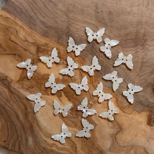 Könnte beinhalten: Eine Sammlung von zwanzig kleinen, weißen, schillernden schmetterlingsförmigen Knöpfen. Jeder Knopf hat eine glatte, perlmuttartige Oberfläche und zwei kleine Löcher in der Mitte des Schmetterlingskörpers zur Befestigung an Stoffen.