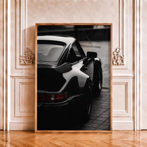 Peut inclure: Une impression d'art murale encadrée présentant une photographie en noir et blanc de la vue arrière trois-quarts d'une voiture de sport classique. Le véhicule présente un aileron arrière proéminent, des lignes courbes et un feu arrière rectangulaire.