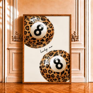 Peut inclure: Une impression d'art encadrée représentant deux boules de billard avec un motif léopard marron et noir. Chaque boule possède un centre circulaire blanc avec un chiffre huit noir. Le texte "lucky me" est écrit en cursive noire entre elles.