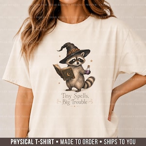 May include: A cream-colored t-shirt featuring a graphic of a raccoon wearing a black witch hat, holding an open book and a small purple potion bottle. Below the raccoon, the text reads Tiny Spells, Big Trouble, surrounded by small stars.