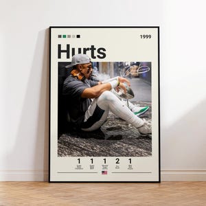 May include: A framed poster featuring a photograph of a football player wearing a grey cap, dark shirt, and white leggings, sitting on the ground while holding a football. The text reads Hurts, 1999, and lists various football achievements below.