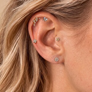 May include: A set of five gold-tone ear piercings featuring a curved leaf-shaped stud with clear stones, a small four-petaled flower stud, a larger flower stud with a yellow centre, a tiny flower stud, and a round floral-shaped stud with clear stones.