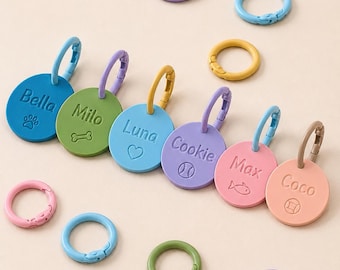 May include: A collection of circular, matte-finish pet identification tags in blue, green, light blue, purple, pink, and peach. Each tag features a name and a small icon, including a paw, bone, heart, baseball, fish, and ball, with matching rings.