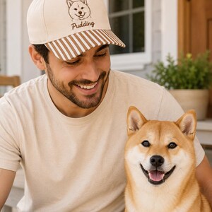 May include: A cream-colored baseball cap featuring a brown embroidered line art illustration of a Shiba Inu dog face above the text "Pudding" in brown script. The cap has a curved brim with a brown and white vertical striped pattern.