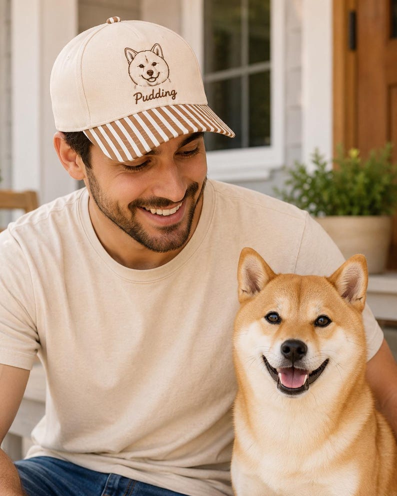 May include: A cream-colored baseball cap featuring a brown embroidered line art illustration of a Shiba Inu dog face above the text "Pudding" in brown script. The cap has a curved brim with a brown and white vertical striped pattern.
