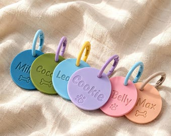 May include: A set of six circular pet tags in blue, olive green, light blue, lavender, pink, and peach. Each tag features a name and a small embossed bone or paw print icon, with a matching colored plastic ring attached to the top of each.