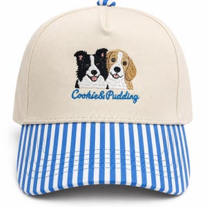May include: A beige baseball cap with a blue and white striped brim and a blue and white striped button on top. The front features an embroidered graphic of a black and white dog and a tan and white dog with the blue text Cookie & Pudding below.