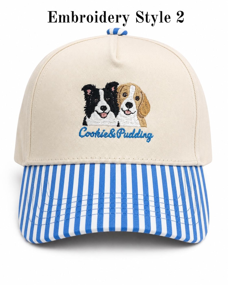 May include: A beige baseball cap with a blue and white striped brim and a blue and white striped button on top. The front features an embroidered graphic of a black and white dog and a tan and white dog with the blue text Cookie & Pudding below.