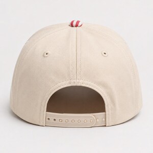 May include: A beige baseball cap featuring a structured crown with six panels and embroidered eyelets. The top of the cap is accented with a small, round button in a red and white striped pattern. An adjustable plastic snapback closure is at the rear.