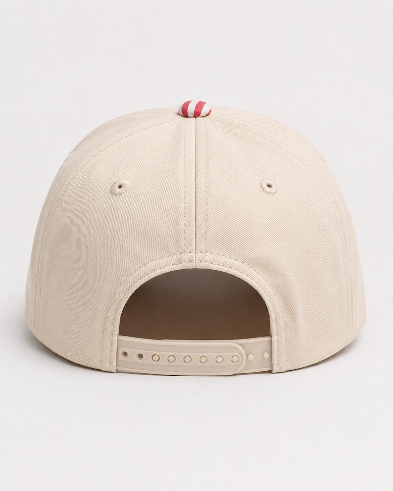 May include: A beige baseball cap featuring a structured crown with six panels and embroidered eyelets. The top of the cap is accented with a small, round button in a red and white striped pattern. An adjustable plastic snapback closure is at the rear.