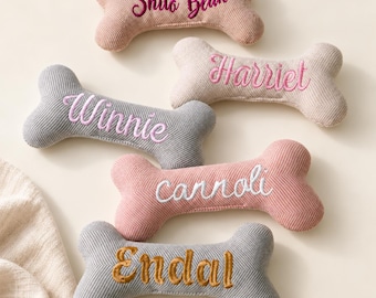 May include: Five bone-shaped fabric toys with embroidered names. The top bone is tan with 'Shilo Bean' in dark pink. Below is a beige bone with 'Harriet' in pink, a gray bone with 'Winnie' in pink, a pink bone with 'cannoli' in white, and a gray bone with 'Endal' in gold.