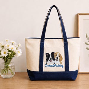Op de afbeelding: Een crèmekleurige canvas tas met marineblauwe randen en hengsels. De voorzijde is voorzien van een borduursel van een zwart-witte hond en een bruin-witte hond, met daaronder de blauwe geborduurde tekst Cookie & Pudding.