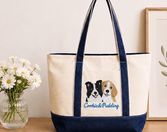 May include: A cream-colored canvas tote bag with navy blue trim and handles. The front features an embroidered graphic of a black and white dog and a tan and white dog, with the blue embroidered text Cookie & Pudding positioned below them.