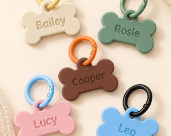 May include: Five bone-shaped pet ID tags in cream, green, brown, pink, and blue, each featuring a matching or contrasting circular clip. The tags are embossed with the names Bailey, Rosie, Cooper, Lucy, and Leo in a simple font.