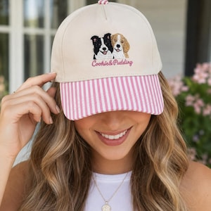 May include: A beige baseball cap featuring an embroidered graphic of a black and white dog next to a tan and white dog. Below the dogs, the text Cookie & Pudding is embroidered in pink. The cap has a pink and white vertical striped brim.