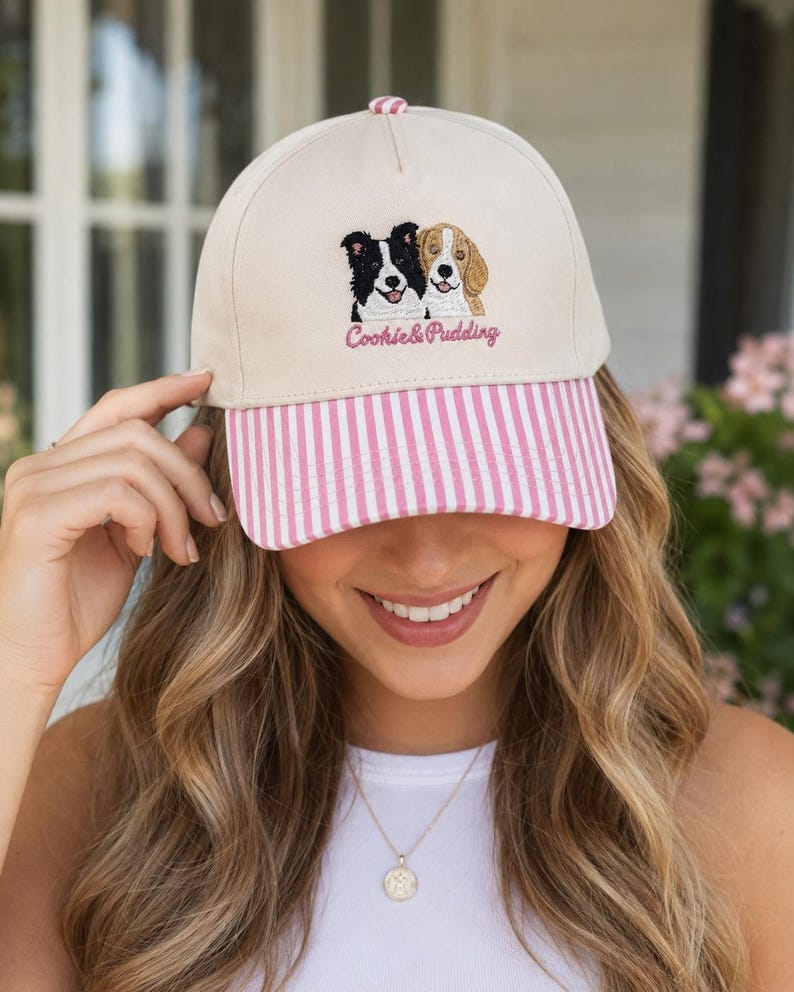 May include: A beige baseball cap featuring an embroidered graphic of a black and white dog next to a tan and white dog. Below the dogs, the text Cookie & Pudding is embroidered in pink. The cap has a pink and white vertical striped brim.