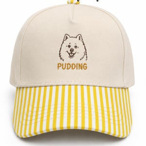May include: A cream-colored baseball cap featuring a brown embroidered dog illustration above the text PUDDING in gold thread. The cap has a yellow and white vertical striped brim and a small yellow and white striped button on the top crown.