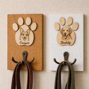 Könnte beinhalten: Zwei rechteckige Wandhaken aus Holz mit lasergravierten Pfotenabdrücken und Hundeporträts. Das linke Brett ist aus Naturholz mit einem Corgi namens Cooper, das rechte ist weiß mit einem Labrador namens Buddy. Jeder hat einen bronzefarbenen Haken.
