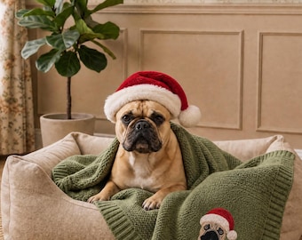 May include: A fawn-colored French Bulldog wearing a red and white plush Santa hat, resting in a beige pet bed while covered by a textured olive green knit blanket that features an embroidered patch of a French Bulldog in a Santa hat.