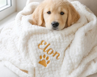 May include: A soft, white, textured blanket wrapped around a golden retriever puppy. The blanket features the name Elon embroidered in gold thread above a gold embroidered paw print. The fabric has a raised, bumpy, plush surface texture.