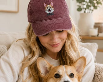 May include: A maroon baseball cap featuring an embroidered corgi dog face on the front panel. Below the dog illustration, the name Milo is stitched in dark green cursive script. The cap has a distressed, washed-out fabric texture.