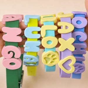 May include: Five silicone pet collars in various colours including green, lime, light blue, lavender, and blue. Each collar features raised, colourful, block-letter names such as MANGO, LUNA, and JAX, along with decorative paw print and heart shapes.