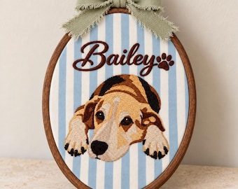 May include: An oval wooden embroidery hoop containing fabric with light blue and white vertical stripes. The embroidery features a brown and tan dog with a black nose and paws, the name Bailey in brown script, and a brown paw print.
