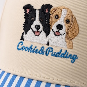 May include: A beige baseball cap featuring an embroidered design of a black and white dog alongside a tan and white dog. Below the dogs, the text Cookie & Pudding is embroidered in blue script. The brim features a blue and white striped pattern.