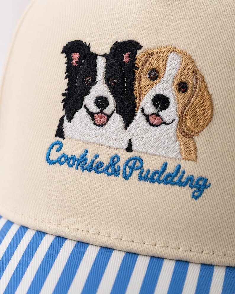 May include: A beige baseball cap featuring an embroidered design of a black and white dog alongside a tan and white dog. Below the dogs, the text Cookie & Pudding is embroidered in blue script. The brim features a blue and white striped pattern.