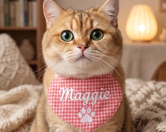 May include: A golden-brown cat with green eyes wearing a heart-shaped pink and white gingham bandana. The bandana features the name Maggie embroidered in white script lettering above a white embroidered paw print icon centered on the fabric.