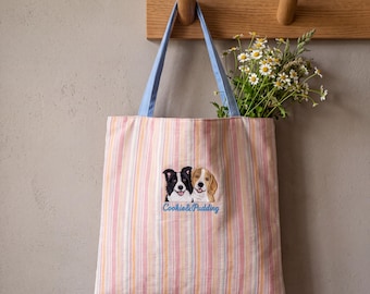 May include: A vertical striped tote bag in shades of pink, orange, and white with light blue handles. The center features an embroidered graphic of a black and white dog and a tan dog with the blue embroidered text Cookie & Pudding below them.
