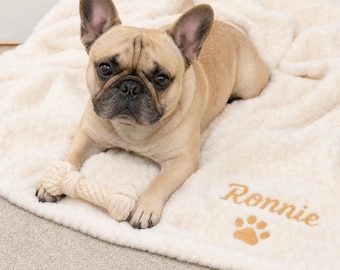 May include: A fawn French Bulldog lying on a cream-colored, textured fleece blanket. The blanket features the embroidered name Ronnie in gold thread, accompanied by a gold paw print icon. A white rope toy rests between the dog's front paws.