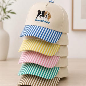 May include: A stack of five baseball caps with cream-colored crowns and embroidered dog faces above the text Cookie & Pudding. Each cap features a different colored brim with vertical stripes in blue, yellow, pink, green, and brown respectively.