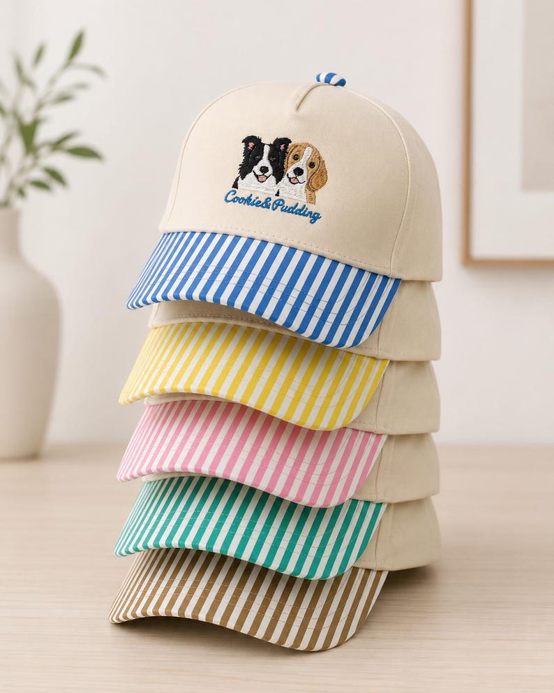 May include: A stack of five baseball caps with cream-colored crowns and embroidered dog faces above the text Cookie & Pudding. Each cap features a different colored brim with vertical stripes in blue, yellow, pink, green, and brown respectively.