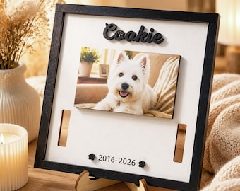 May include: A square black wooden frame with a white backing featuring a centered rectangular photo of a white dog. The name Coakie is in black script above the photo, and the dates 2016-2026 are printed below with two small black paw prints.