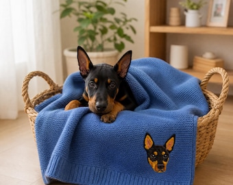 May include: A blue knit blanket featuring an embroidered portrait of a black and tan dog. The blanket is draped inside a woven wicker basket, with a black and tan dog resting on top of the fabric, partially covered by the blue knit material.