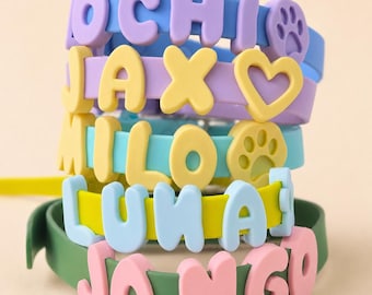 May include: A stack of five silicone pet collars in blue, purple, teal, yellow, and green. Each collar features raised, colorful, block-letter charms spelling out the names OCHI, JAX, MILO, LUNA, and JANGO, along with heart and paw print icons.