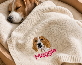 May include: A cream-colored knit blanket featuring an embroidered beagle portrait and the name Maggie stitched in bright pink thread. The blanket is draped over a sleeping beagle dog resting inside a round wooden pet bed with a soft cushion.