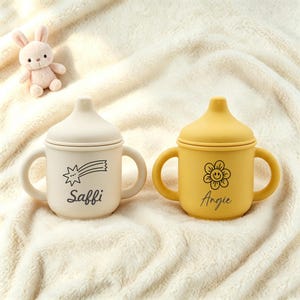 Può includere: Due tazze con beccuccio in silicone dotate di due manici e coperchio. La tazza a sinistra è color crema con una stella cadente nera e il nome Saffi. La tazza a destra è gialla con un fiore sorridente nero e il nome Angie stampato sotto.