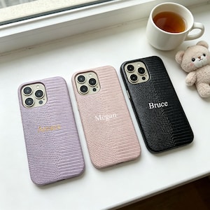 Può includere: Tre cover per smartphone con una texture a pelle di lucertola nei colori lavanda, rosa chiaro e nero. La cover lavanda riporta il nome Amara, quella rosa il nome Megan e la cover nera riporta il nome Bruce in caratteri bianchi.