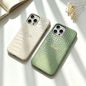 Può includere: Due cover per smartphone con una texture a motivo pelle di coccodrillo. Una cover è color crema con il nome Cyra stampato al centro, l'altra è verde salvia con il nome Beah stampato in lettere dorate al centro del retro della cover.